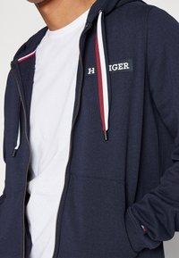 Felpa con zip color navy realizzata in tessuto morbido, dotata di tasca frontale, con cordini bianchi, rossi e blu e un patch con logo sul petto.