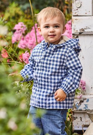 Blaue und weiße Karojacke mit Kapuze, elastischen Bündchen, kombiniert mit Jeans. Umgeben von pinken Blumen und Grünpflanzen.