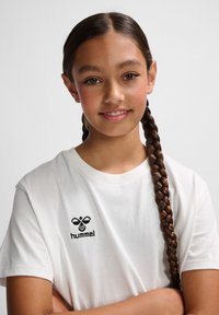 Hvid bomulds-t-shirt med sort broderet logo på brystet. Modellen har langt fletet hår, armene krydset, og smiler imod en lys baggrund.