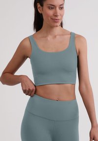 Reggiseno sportivo verde acqua con spalline spesse, scollatura arrotondata e texture liscia, indossato con leggings coordinati che mettono in mostra un design aderente.