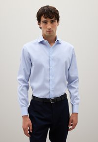 Hombre vestido con una camisa de botones a rayas azul claro y blanca, metida dentro de pantalones oscuros de color navy con un cinturón negro, de pie frente a un fondo liso.