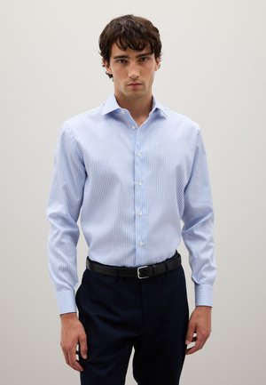 Hombre vestido con una camisa de botones a rayas azul claro y blanca, metida dentro de pantalones oscuros de color navy con un cinturón negro, de pie frente a un fondo liso.