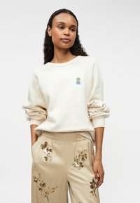 Krém színű pulóver bordázott mandzsettákkal, a bal mellrészen egy kicsi, színes logóval. Bézs nadrággal párosítva, amelyet arany flitteres virágminták díszítenek.