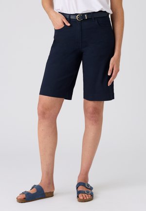 Mujer con pantalones cortos azul marino hasta la rodilla con cinturón negro, top blanco y sandalias azules con hebilla, de pie frente a un fondo liso.