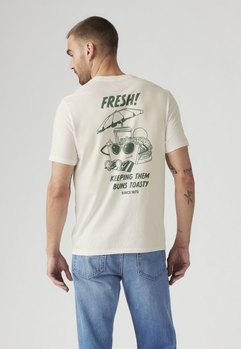 Άνδρας που φοράει λευκό T-shirt με καρτούν ποτό, μπέργκερ και κείμενο "FRESH! Keeping them buns toasty since 1873" στην πλάτη, σε συνδυασμό με μπλε τζιν.