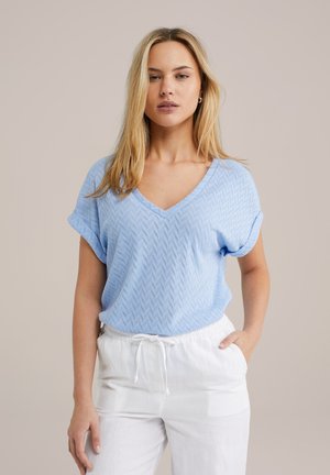 Light blue, short-sleeve V-neck top with a zigzag pattern; worn with white, drawstring pants. Smooth texture, relaxed fit.