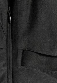 Gros plan sur un tissu noir avec une fermeture éclair verticale et des panneaux textiles superposés cousus pour un design structuré.