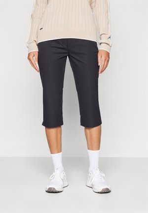 Person iført beige ribstrikket sweater, sorte korte bukser der ender under knæet, hvide crew-sokker og hvide sneakers, stående på gråt gulv.