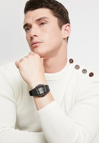 Digital watch with a black resin strap and square face, featuring an LCD screen with red and grey accents. White textured sweater with buttons.
