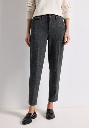 Personne portant un pantalon gris foncé sur mesure avec un motif à fines carreaux, des mocassins noirs et un pull crème, debout devant un fond clair uni.