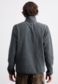 Γκρι φλις πόλο με υψηλό γιακά και μακριά μανίκια. Μαλακή υφή, χαλαρή εφαρμογή. Συνδυάζεται με καφέ cargo παντελόνι.