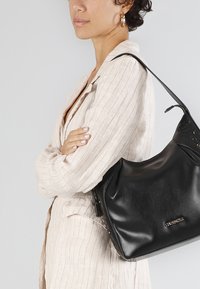 Borsa a spalla in pelle nera con una texture liscia, forma curvata e logo in tonalità oro. Presenta dettagli perforati e una sola tracolla.