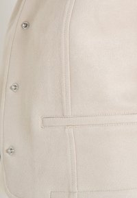 Gros plan sur l'avant d'une veste en daim beige avec des boutons-pression argentés et des détails de poches passepoilées horizontales.