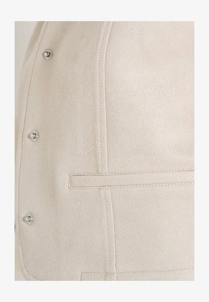 Gros plan sur l'avant d'une veste en daim beige avec des boutons-pression argentés et des détails de poches passepoilées horizontales.