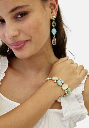 Mujer que lleva pendientes colgantes con piedras preciosas multicolores y pulsera de oro a juego con piedras en tonos pastel y transparentes, vestida con una blusa blanca sin mangas con volantes.