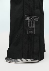 Μαύρο παντελόνι Adidas με κουμπιά πατιλέτας και επώνυμα μπαλώματα που καλύπτουν μερικώς ασημένια και λευκά αθλητικά παπούτσια Adidas.