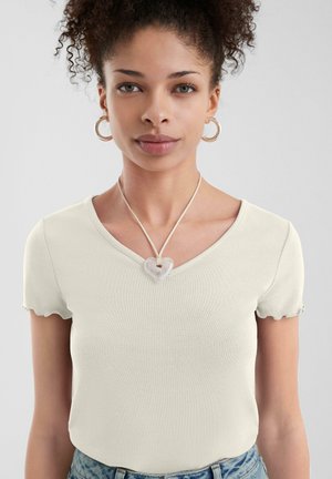 Top a maniche corte, di un beige chiaro, con scollatura a V e bordi ondulati, abbinato a una collana con ciondolo a forma di cuore trasparente. Sono visibili jeans in denim blu.