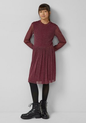 Bordeauxfarbenes langärmeliges Kleid mit einem plissierten Rock, gefertigt aus einem glitzernden Stoff. Schwarze Stiefeletten ergänzen das Outfit, getragen mit schwarzen Strumpfhosen.