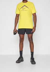 Keltainen Nike Trail -t-paita, jossa on graafinen logo, yhdistettynä mustiin urheilushortseihin, mustiin sukkiin ja harmaisiin urheilukenkiin. Pitää kädessä mustaa juomapulloa.