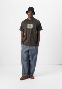 T-shirt marron foncé avec un design graphique coloré et du texte, associé à un pantalon en denim bleu ample et un chapeau seau noir. Chaussures marron.