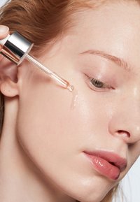 Goccia di siero trasparente applicata sulla guancia utilizzando un contagocce di vetro tenuto vicino al viso con pelle chiara e trucco naturale.