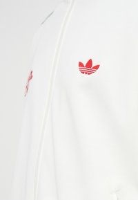 Weißes Sweatshirt mit Reißverschluss, das ein gesticktes rotes Drei-Streifen-Logo auf der linken Brust und eine subtile Textur im Stoff aufweist.