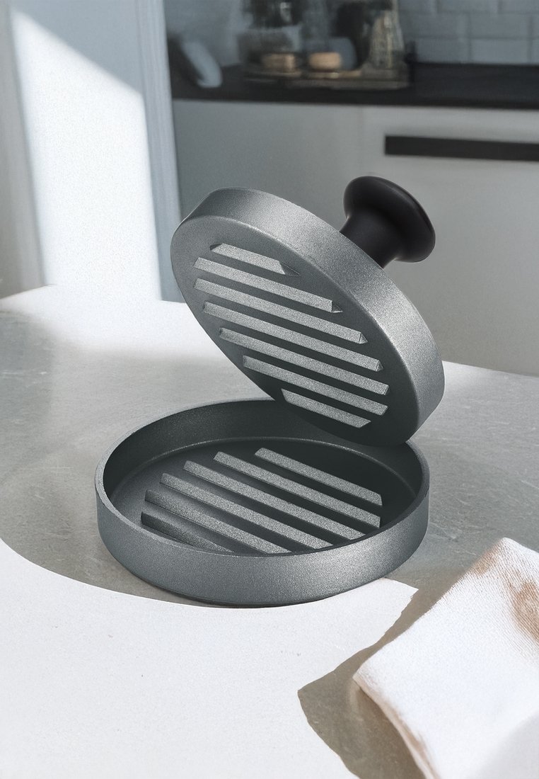 Wyciskacz do burgerów z żeliwa, szary kolor, okrągły kształt, karbowane wnętrze, ergonomiczny czarny uchwyt, umieszczony na jasnym blacie kuchennym z ręcznikiem w pobliżu.
