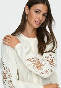 Biała bluzka zapinana na guziki z długimi, obszernymi rękawami, zdobiona misterną koronką i o teksturowanym wykończeniu. Miękka, lekka tkanina.