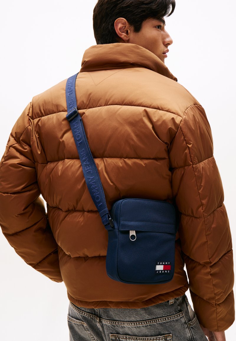 Giacca a imbottitura marrone con colletto alto, borsa a tracolla blu navy con chiusura a zip e toppa con logo, indossata sopra jeans sbiaditi.