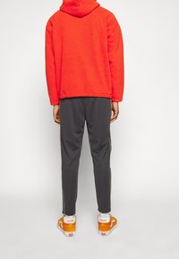 Person som bär en röd fleecehuva, mörkgrå träningsoverallsbyxor med sidostråk, och orange Vans sneakers, står med ryggen mot en vit bakgrund.