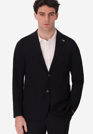 Uomo che indossa una giacca da completo nera sopra una camicia bianca con colletto, in piedi con una mano in tasca contro uno sfondo uniforme.