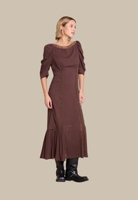 Brun knelang kjole med pufferm, blondehals, og vertikale sømmer. Har volangkant og intrikate detaljer. Svarte knelange støvler.