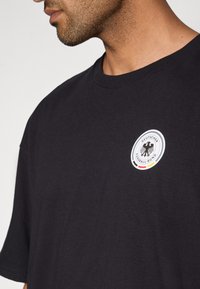 Fekete pamut póló kerek nyakkivágással. Közepén egy kör alakú fehér folt látható fekete sas ábrával és a "DEUTSCHER FUSSBALL-BUND" felirattal.