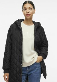 Črna prešita jakna s kapuco, opremljena z naprstniki in elastičnimi manšetami, nosi se preko svetlo bež puloverja s tiskom "Razišči".