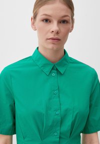 Zielona koszula z krótkim rękawem, z wykładanym kołnierzykiem i zapięciem na guziki. Wykonana z gładkiego materiału o lekko teksturowanym wykończeniu.
