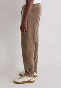 Person, der beiges Cord-Hosen und weiße Turnschuhe trägt und seitlich an einer schlichten hellen Wand steht.