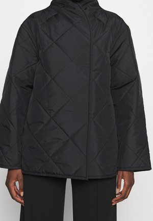 Veste matelassée noire avec surpiqûres en losange, manches longues et col montant, portée sur un pantalon noir.