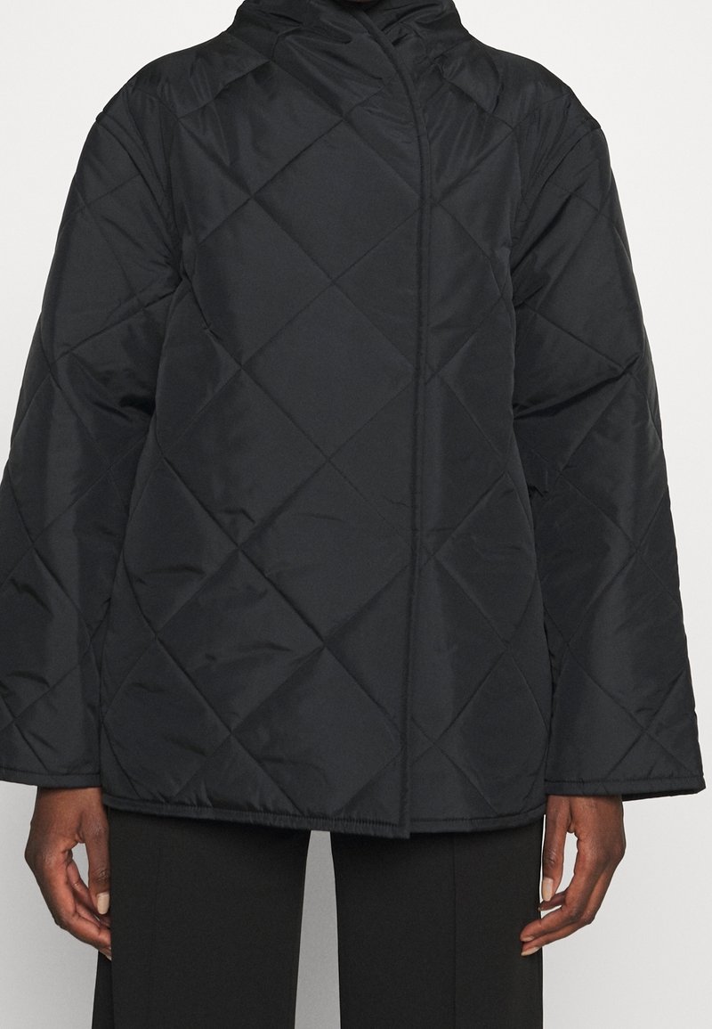 Veste matelassée noire avec surpiqûres en losange, manches longues et col montant, portée sur un pantalon noir.