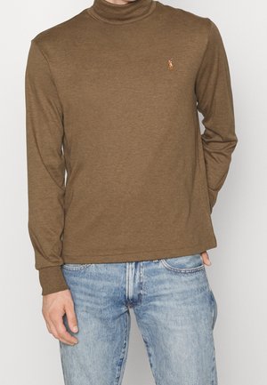 Mann trägt einen braunen Langarm-Rollkragenpullover mit kleinem gesticktem Logo auf der Brust und hellblaue Jeans.