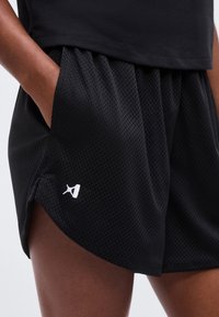 Άτομο φοράει μαύρα αθλητικά σορτς από διάτρητο ύφασμα με μικρό λευκό λογότυπο στο αριστερό τελείωμα, χέρι στην τσέπη, σε συνδυασμό με μαύρο τοπ.