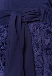 Navy-asuste, jossa on kangasvyö ja kukkakirjailtu pinta. Sujuva yläosa, jossa on rypistetty vyötärö, esittelee monimutkaisia kuvioita tummansinisessä.