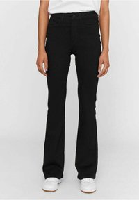 Personne portant un pantalon noir taille haute à jambes droites, des baskets blanches et une chemise blanche, debout devant un fond uni.