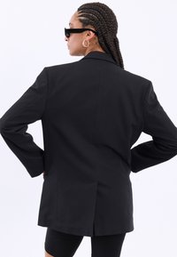 Schwarzer, taillierter Blazer mit einem einzelnen Rückenschlitz, strukturierten Schultern und langen Ärmeln. Glattes Gewebe, keine sichtbaren Muster oder Hardware-Details.