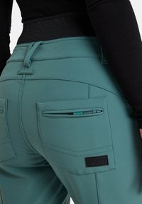 Türkisfarbene, hochgeschnittene Hose mit glatter Textur, ausgestattet mit hinteren Reißverschlusstaschen und einem Logopatch für zusätzliche Details.