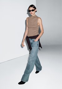 Brown sleeveless turtleneck, dark sweater draped at waist, light blue flared jeans, and black pointed-toe high heels. Solid color and minimal design.