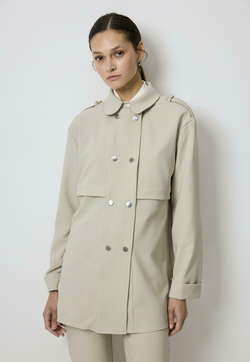 Kobieta o ciemnych włosach związanych do tyłu, ubrana w beżową dwurzędową marynarkę z srebrnymi guzikami oraz beżowe spodnie, stojąca na białym tle.