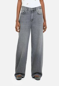 Person wearing high-waisted, loose-fitting gray jeans and black pointed flats with a textured pattern, standing against a white background.