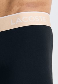 Tissu extensible noir avec une ceinture élastique beige, arborant le texte blanc "LACOSTE". Le design est simple et ajusté.