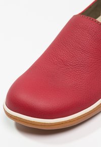 Chaussure en cuir rouge à enfiler avec une finition texturée, un bout arrondi, des accents blancs le long de la semelle, et une semelle extérieure en caoutchouc beige.