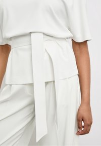 Blusa bianca con cintura legata in vita. Maniche corte e vestibilità comoda con una consistenza liscia, abbinata a pantaloni bianchi larghi.